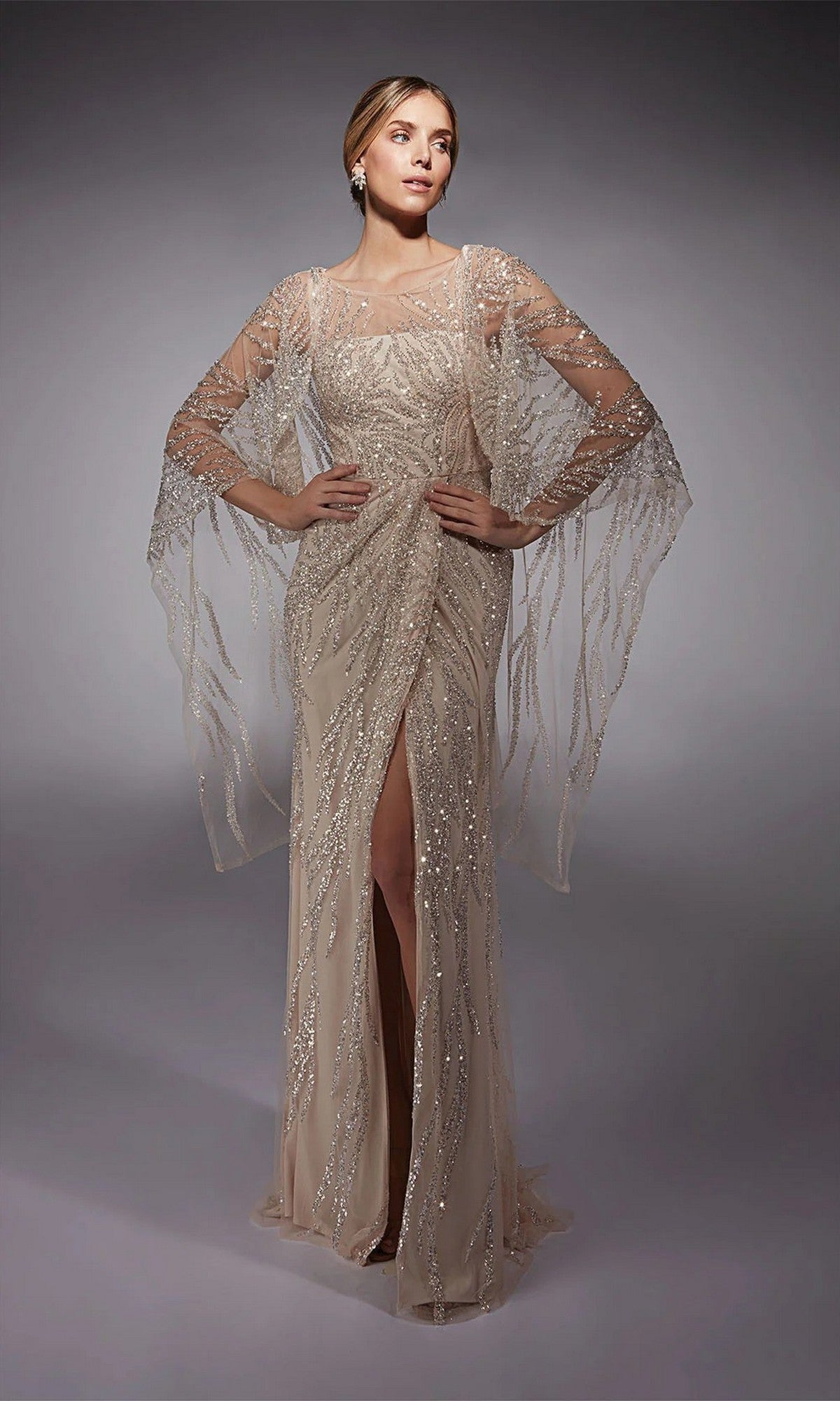 A woman poses in the Alyce 27754, a floor-length beige formal gown with sheer embellished sleeves and a front slit—an elegant mother-of-the-bride choice.